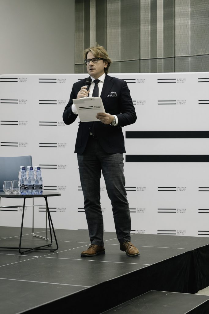 Prof. Wojciech Śleszyński stoi na scenie. W ręku trzyma mikrofon. Przemawia do zgromadzonych osób. W tle za nim jest widoczne logo Muzeum.