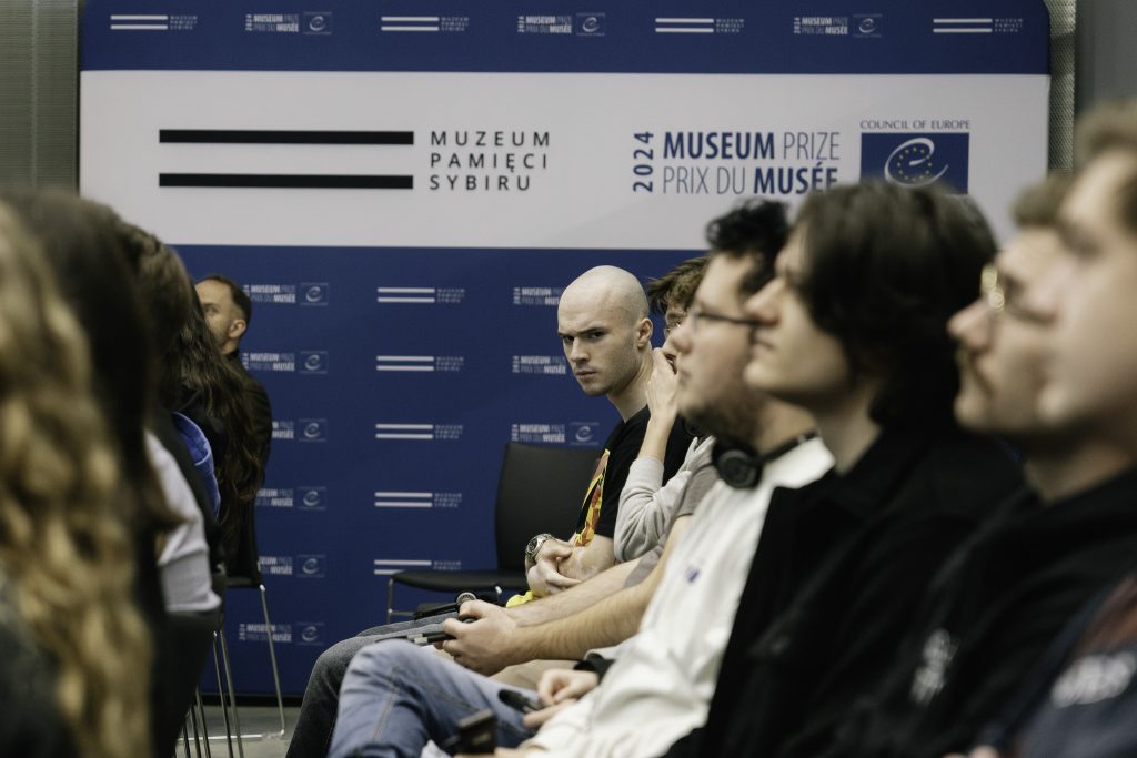 Młodzież w skupieniu słucha przemówienia. W tle widoczna niebieska ścianka z logo Muzeum.