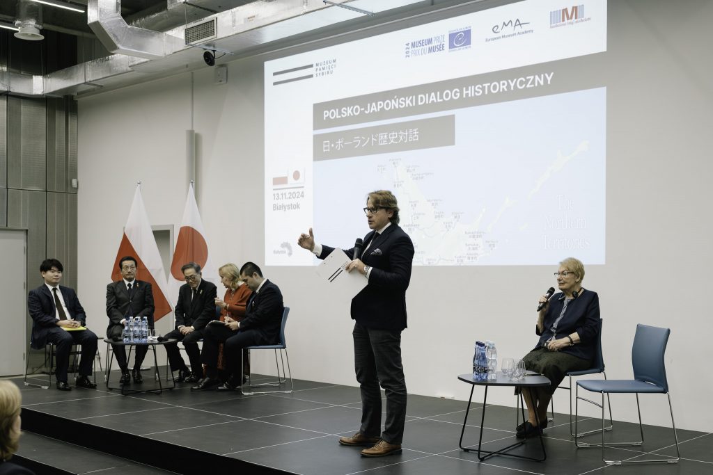 Na scenie są obecni goście z Japonii, dyrektor Muzeum prof. Wojciech Śleszyński, Jolanta Hryniewicka, prezes białostockiego oddziału Związku Sybiraków. Goście z Japonii siedzą przy jednym z dwóch stolików. Przy drugim stoliku siedzi Jolanta Hryniewicka oraz jest jedno wolne miejsce. Na stolikach stoją butelki z wodą oraz szklanki. Dyrektor stoi i przemawia do mikrofonu. W tle za nim jest widoczny slajd z rzutnika, przedstawia wycinek mapy północnej Japonii oraz napis Polsko-Japoński dialog historyczny.
