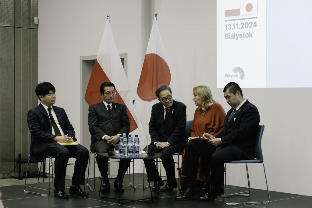 Goście z Japonii siedzą przy jednym z dwóch stolików na scenie. Na stoliku stoją butelki z wodą oraz szklanki. 
