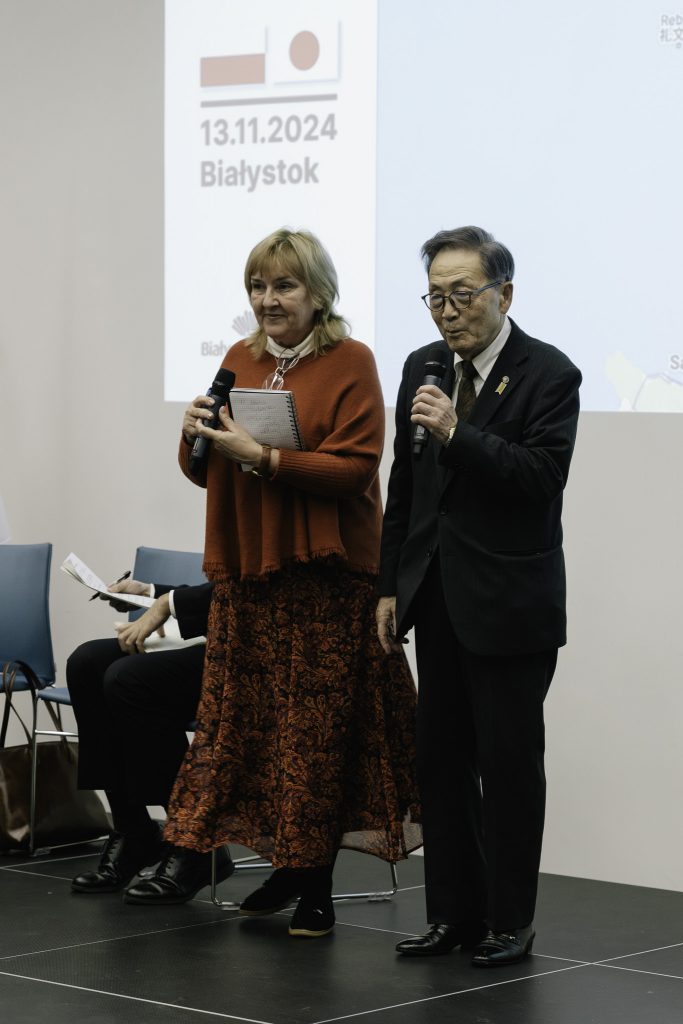 Prezes Matsumoto Yuzo podczas przemówienia. W ręku trzyma mikrofon. Obok niego stoi tłumaczka.