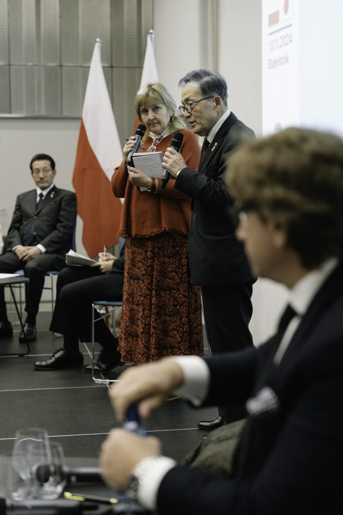 Prezes Matsumoto Yuzo podczas przemówienia. W ręku trzyma mikrofon. Obok niego stoi tłumaczka. W tle widać sylwetki innych osób siedzących na scenie.