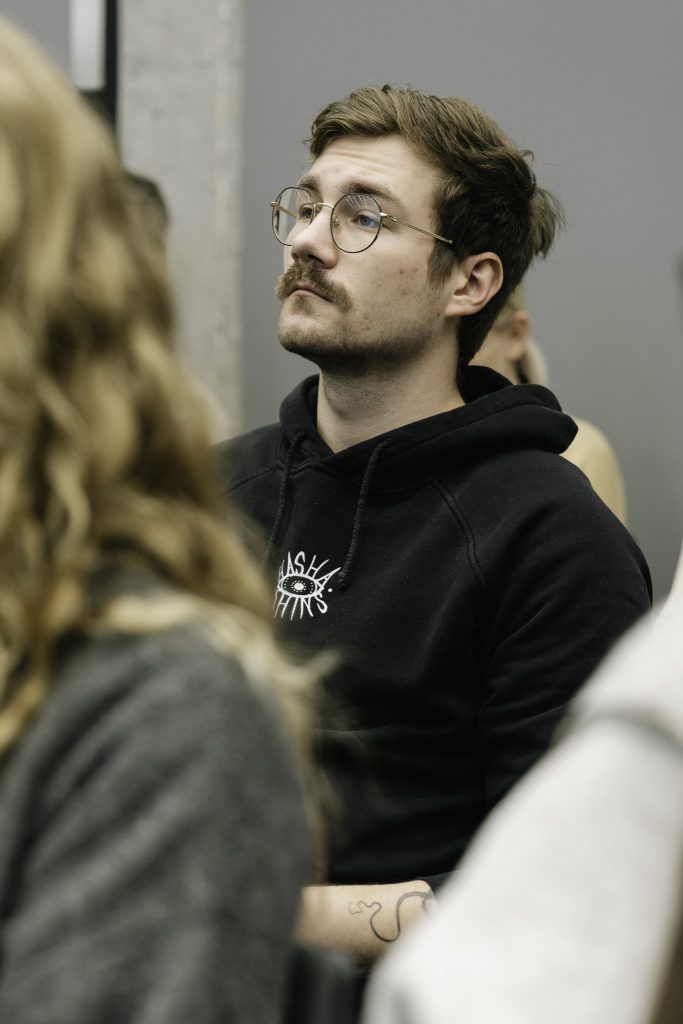 Młody mężczyzna w okularach w skupieniu słucha przemówienia. Jest ubrany w czarną bluzę.