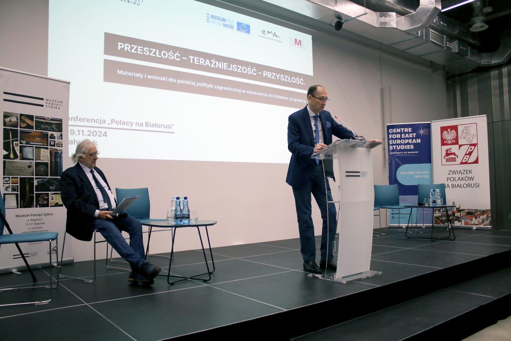 Jeden z uczestników konferencji przemawia przy mównicy. Za jego plecami jest wyświetlany slajd z informacją o konferencji.