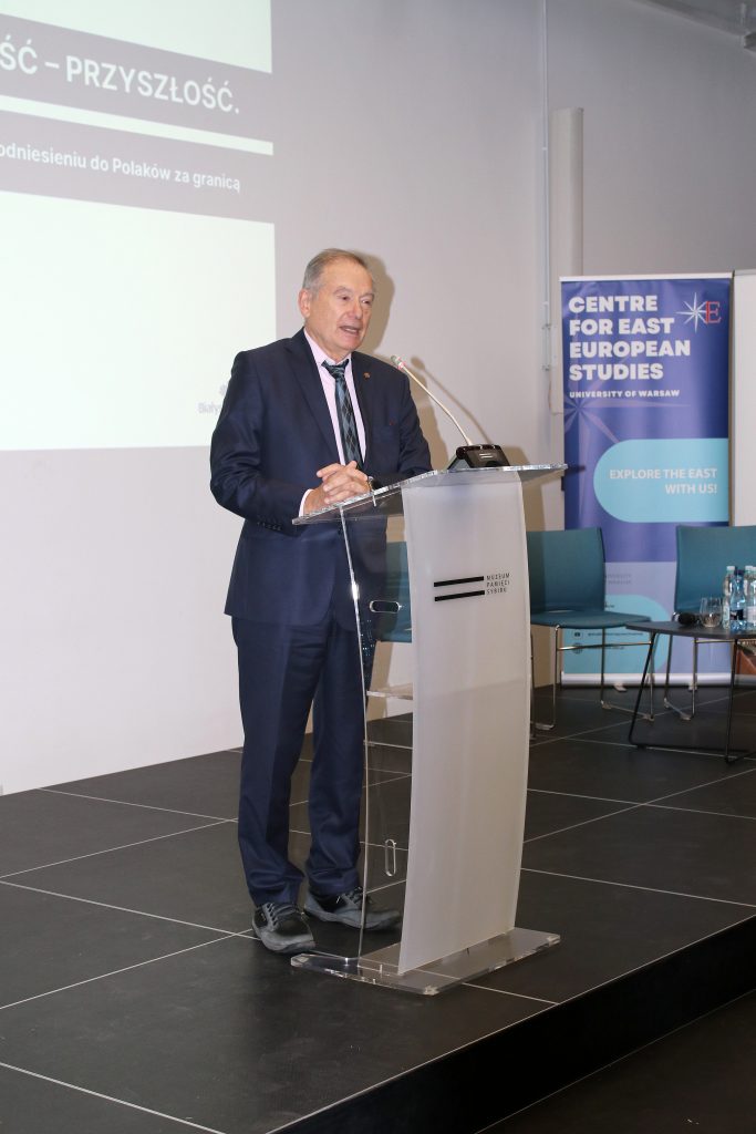 Jeden z uczestników konferencji przemawia przy mównicy. Za jego plecami jest wyświetlany slajd z informacją o konferencji.