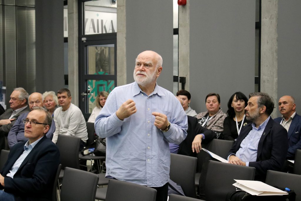 Jeden z uczestników konferencji wypowiada się w dyskusji. Stoi między krzesłami. Inni uczestnicy siedzą i słuchają go.