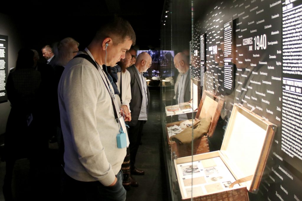 Grupa kilkunastu osób zwiedza wystawę stałą. Stoją przy różnych eksponatach. Część osób ma na uszach słuchawki z audioprzewodnikiem.