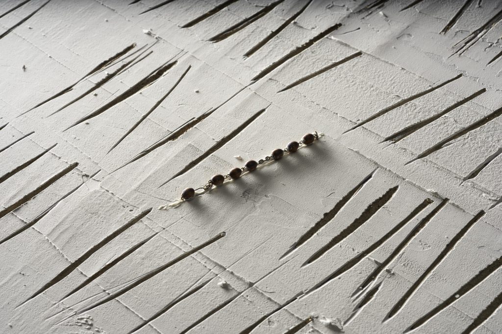 Fragment wystawy czasowej. W betonie jest wyżłobionych wiele pionowych kresek. W centralnym miejscu wisi fragment różańca.