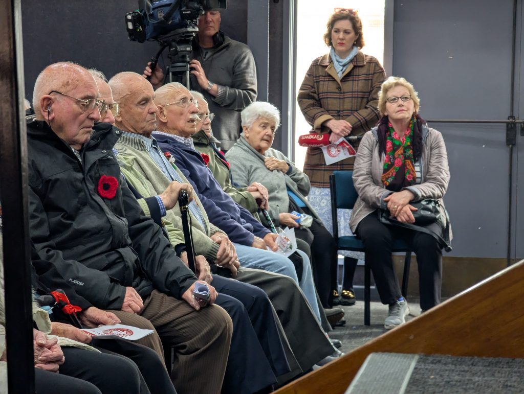 Grupa starszych osób, Sybiraków siedzi na krzesłach. Są ciepło ubrani.