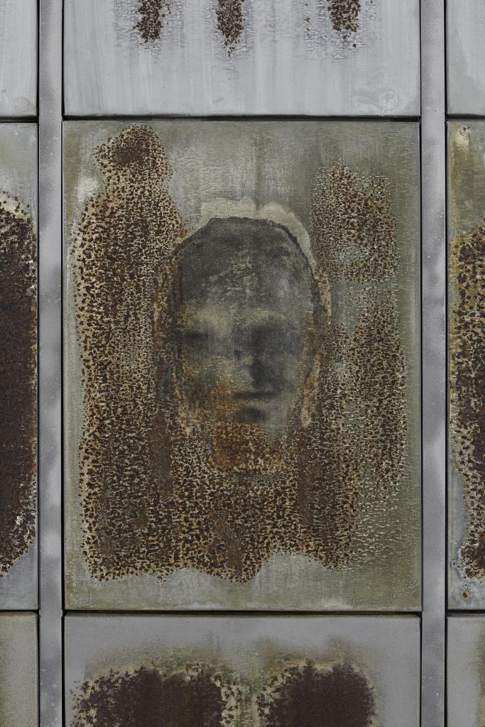 Ciemna, szara, zamazana twarz widniejąca na zardzewiałym arkuszu blachy.