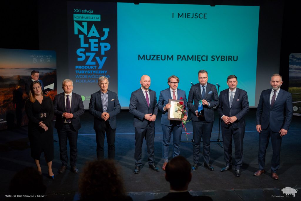 Grupa elegancko ubranych osób stoi na scenie. Wśród nich jest prof. Wojciech Śleszyński, dyrektor Muzeum oraz zastępca prezydenta miasta Białegostoku, Rafał Rudnicki. Dyrektor trzyma w rękach certyfikat oraz różę.  Zastępca prezydenta miasta Białegostoku trzyma w rękach statuetkę.  W tle na dużym ekranie jest wyświetlany napis 1 miejsce Muzeum Pamięci Sybiru.