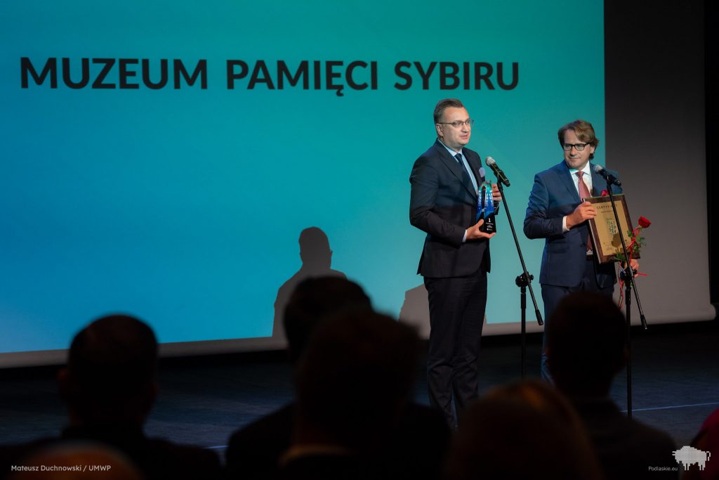 Prof. Wojciech Śleszyński, dyrektor Muzeum oraz zastępca prezydenta miasta Białegostoku, Rafał Rudnicki stoją przy mikrofonach. Dyrektor trzyma w rękach certyfikat oraz różę.  Zastępca prezydenta miasta Białegostoku trzyma w rękach statuetkę.  W tle na dużym ekranie jest wyświetlany napis Muzeum Pamięci Sybiru.
