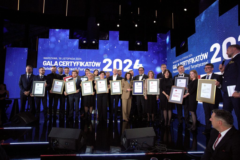 Grupa finalistów konkursu. Wszyscy są elegancko ubrani. W rękach trzymają duże ramki z certyfikatami. W tle jest napis "Gala certyfikatów Polskiej Organizacji Turystycznej".