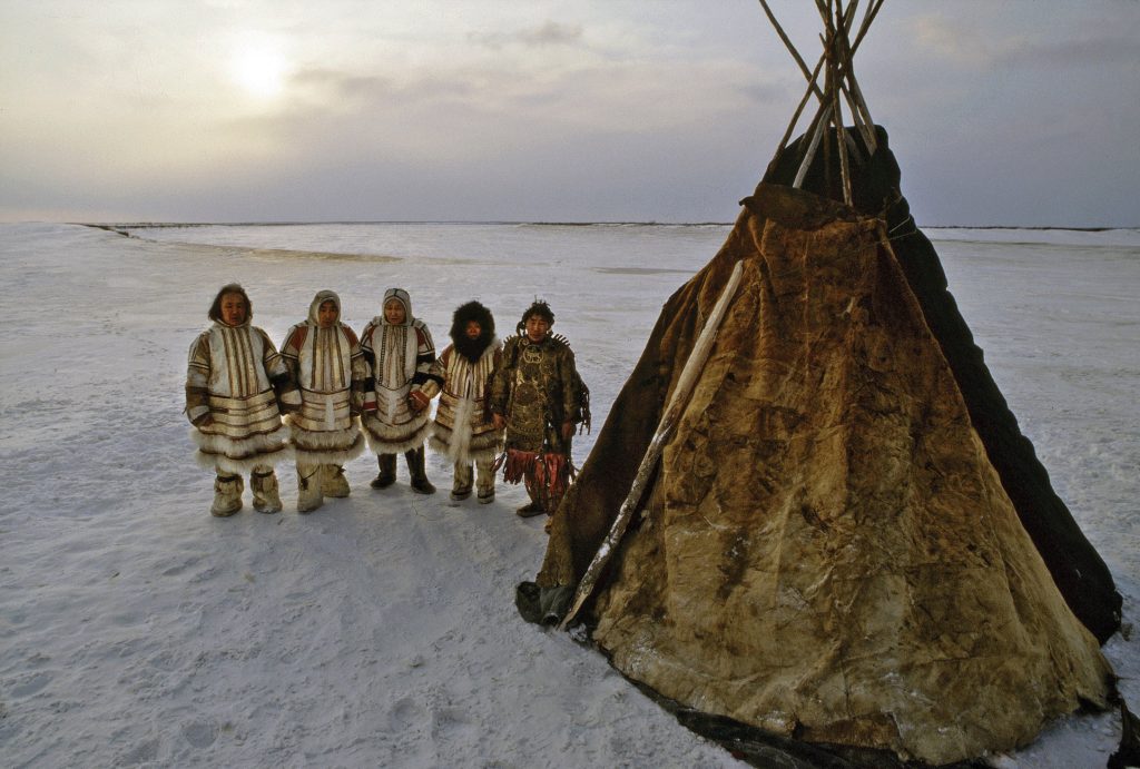 Pięcioro starszych, niewysokich osób w tradycyjnych ubiorach syberyjskich. Ich rysy wskazują że to mogą być rdzenni mieszkańcy regionu. Stoją obok tradycyjnego namiotu pokrytego futrami. Ziemia jest skuta lodem i śniegiem.