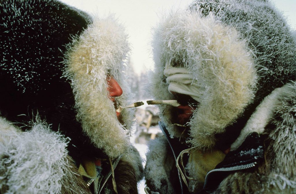 Dwóch mężczyzn w tradycyjnych, ciepłych, futrzanych ubiorach. Jeden z nich odpala swego papierosa od papierosa drugiego mężczyzny. Sceneria sugeruje że jest bardzo zimno.