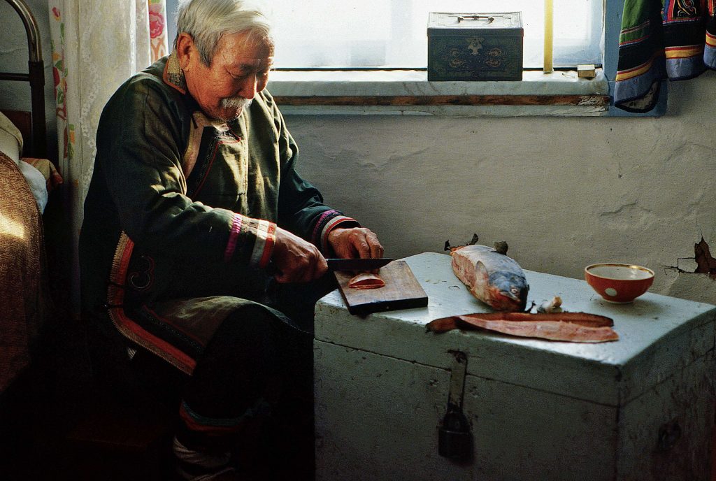 Starszy mężczyzna w tradycyjnym, kolorowym stroju kroi rybę na drewnianej desce. Siedzi przy kufrze w prostym, wiejskim wnętrzu z oknem i łóżkiem.