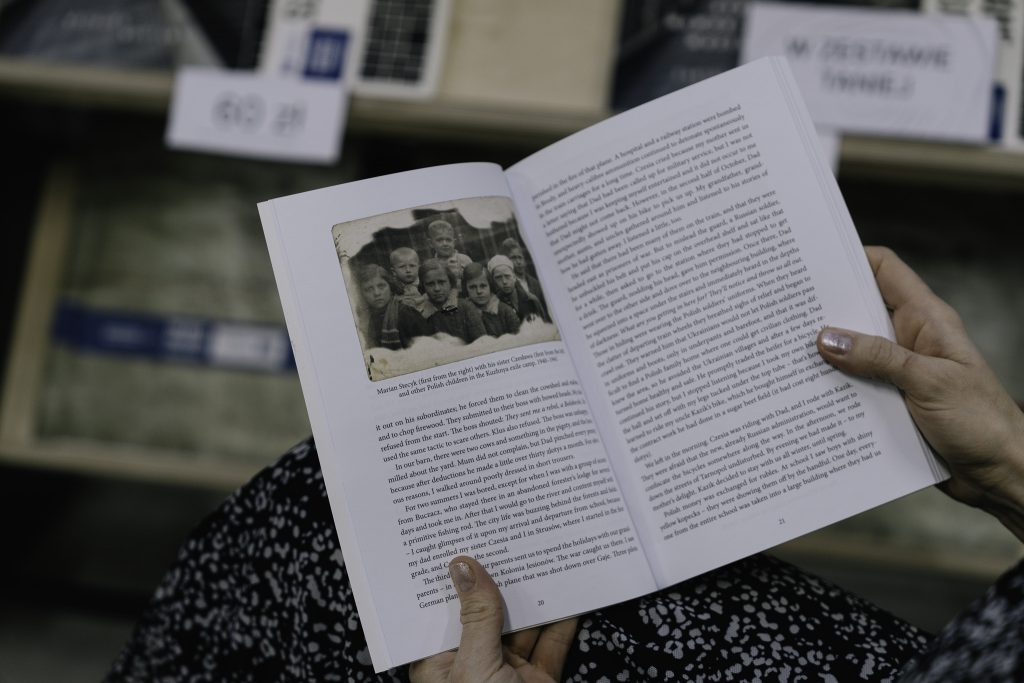 Osoba w czarno-białej spódnicy czyta książkę, trzymając otwartą stronę z czarno-białym zdjęciem grupy dzieci i tekstem.