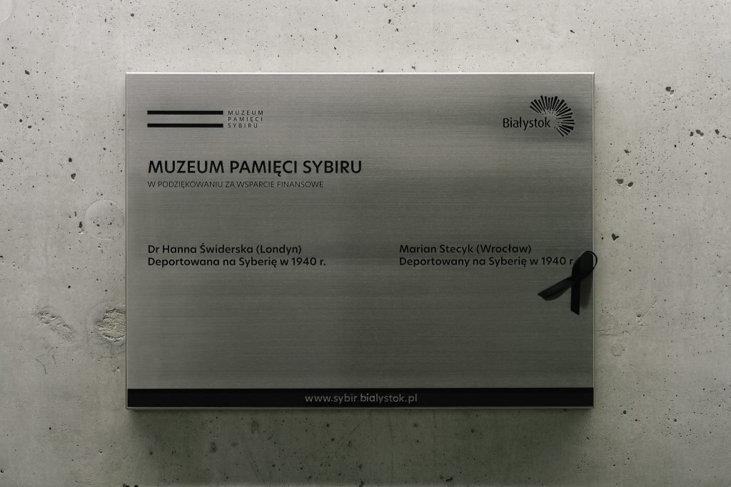 Metalowa tabliczka z napisem "w podziękowaniu za wsparcie finansowe" na tle betonowej ściany. Wymieniono dwie osoby: dr Hannę Świderek i Mariana Stecyka. Obok nazwiska Mariana Stecyka znajduje się czarna wstążka.