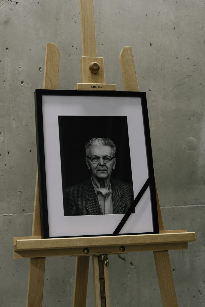 Ramka z czarno-białym zdjęciem Mariana Stecyka umieszczona na drewnianej sztaludze na tle betonowej ściany.