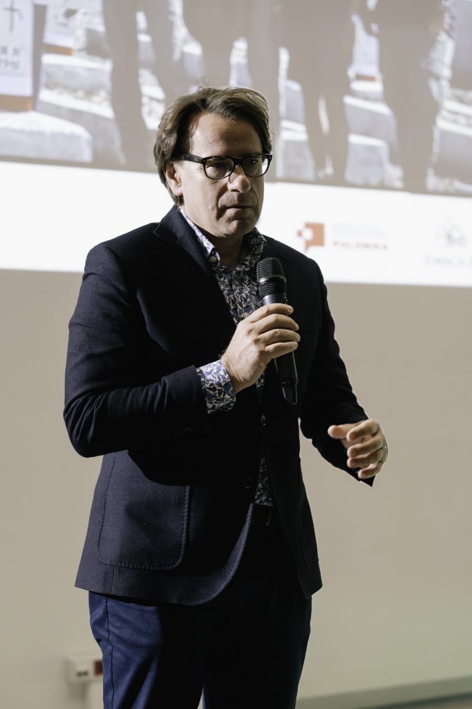 Prof. dr hab. Wojciech Śleszyński przemawia. W ręku trzyma mikrofon. W tle na ścianie jest wyświetlany fragment prezentacji.