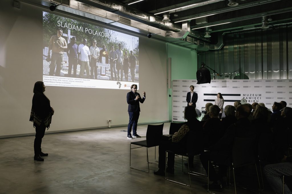 Prof. dr hab. Wojciech Śleszyński przemawia do zgromadzonej publiczności. W ręku trzyma mikrofon. W tle na ścianie jest wyświetlany fragment prezentacji.