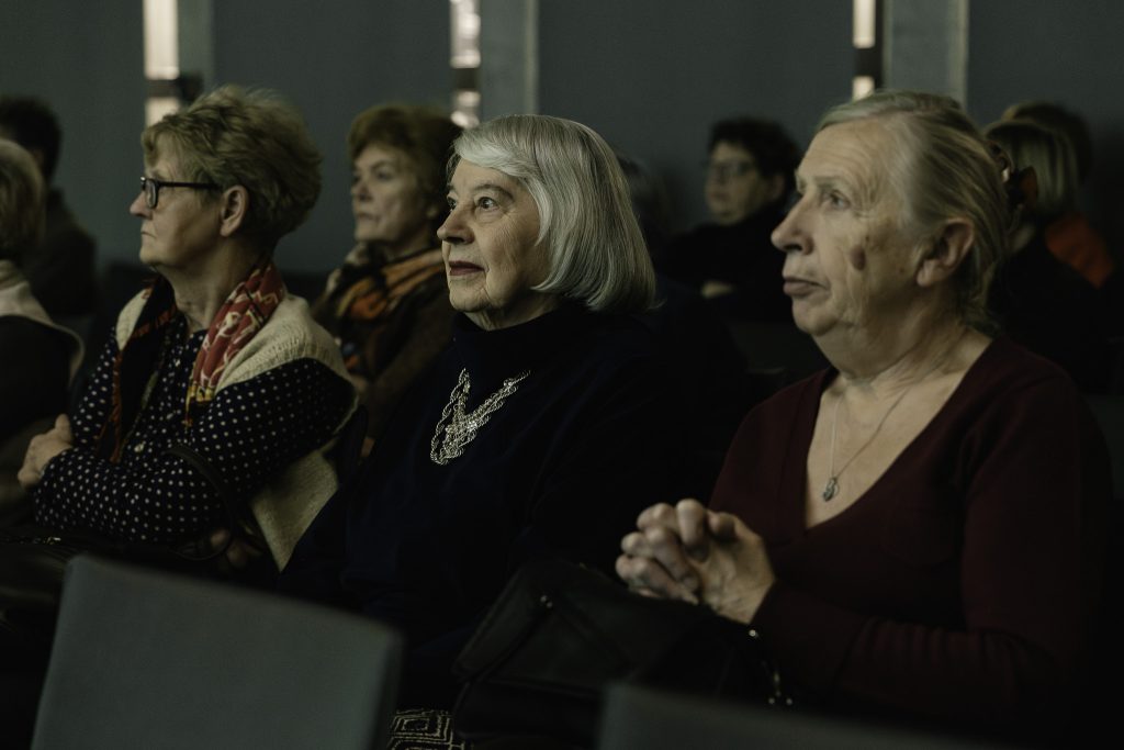 Starsze kobiety siedzące na widowni słuchają w wielkim skupieniu.