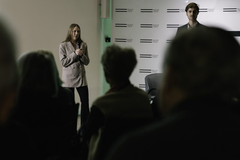 Natalia Mika przemawia. Trzyma w rękach mikrofon. Obok niej stoi Paweł Gniado. W tle widoczna ścianka z logo Muzeum.