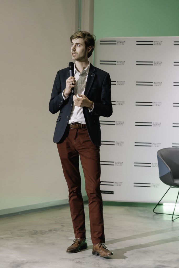 Paweł Gniado przemawia. Trzyma w rękach mikrofon. Patrzy w stronę ekranu z wyświetlaną prezentacją. W tle widoczna ścianka z logo Muzeum.