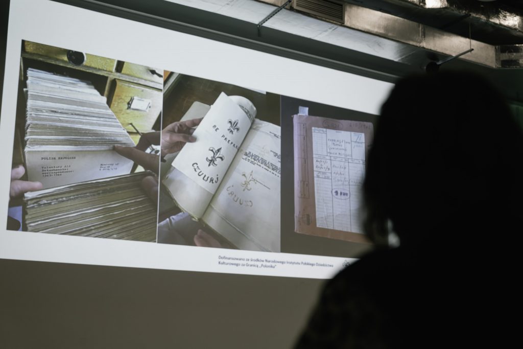 Na dużej ścianie jest wyświetlana prezentacja. Kadr prezentacji pokazuje trzy zdjęcia archiwalnych dokumentów.