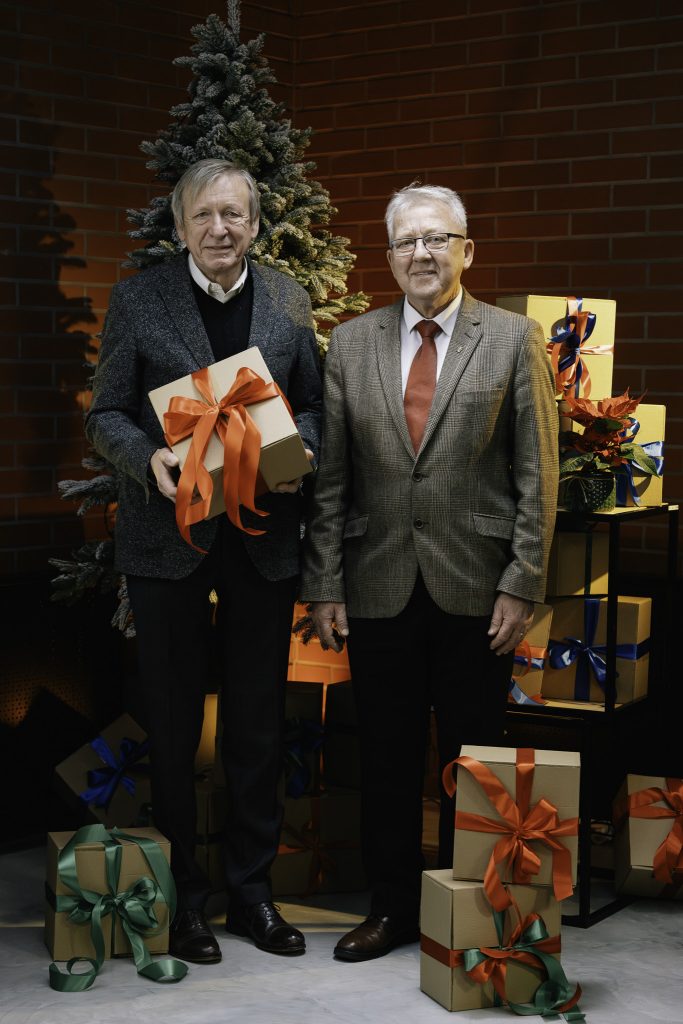 Uśmiechnięci dwaj starsi mężczyźni pozują na tle choinki. Jeden z nich trzyma w rękach prezent z czerwoną wstążką. Wokół nich znajdują się inne prezenty.