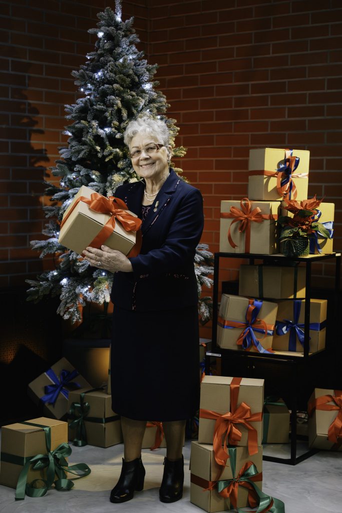 Uśmiechnięta starsza kobieta pozuje na tle choinki, trzymając w rękach prezent z czerwoną wstążką. Wokół niej znajdują się inne prezenty.