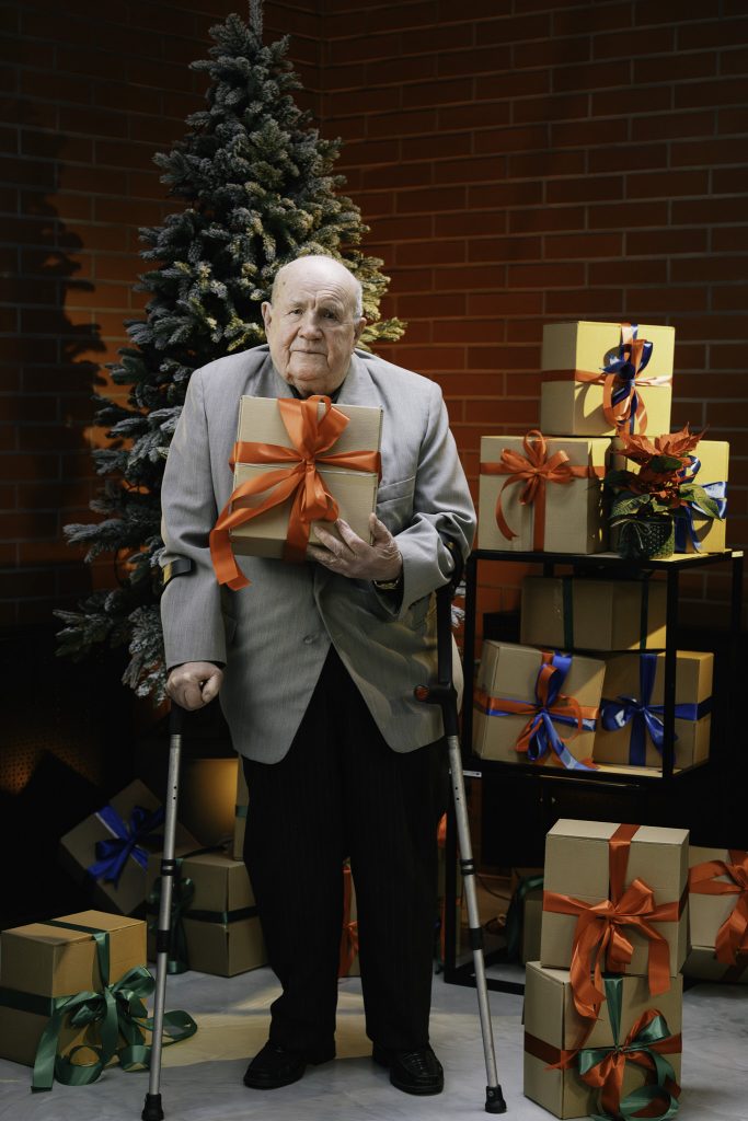 Starszy mężczyzna podparty laskami pozuje na tle choinki, trzymając w rękach prezent z czerwoną wstążką.