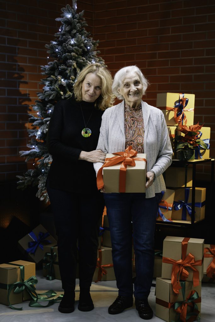 Na tle choinki i prezentów stoją dwie kobiety. Starsza, siwowłosa kobieta uśmiecha się radośnie, trzyma w rękach paczkę. Paczka jest związana dużą czerwoną kokardą. Obok niej stoi kobieta w średnim wieku, uśmiecha się pogodnie.