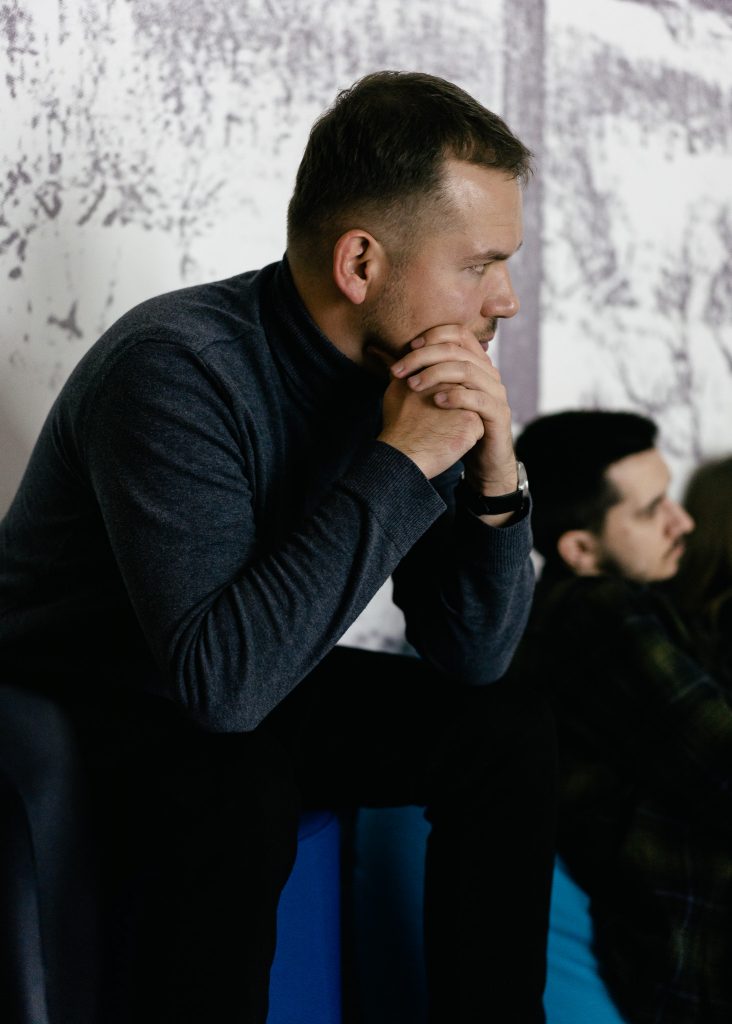 Mężczyzna w szarym golfie siedzi z poważnym wyrazem twarzy, opierając dłonie na brodzie, w jasnym pomieszczeniu. W tle widoczne są rozmyte sylwetki innych osób.