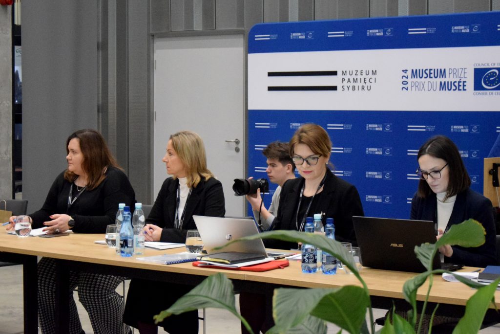 Cztery kobiety w formalnych strojach siedzą przy stole, pracując na laptopach. Na stole znajdują się butelki wody, notatki. W tle jest niebieska ścianka z logo Muzeum Pamięci Sybiru.