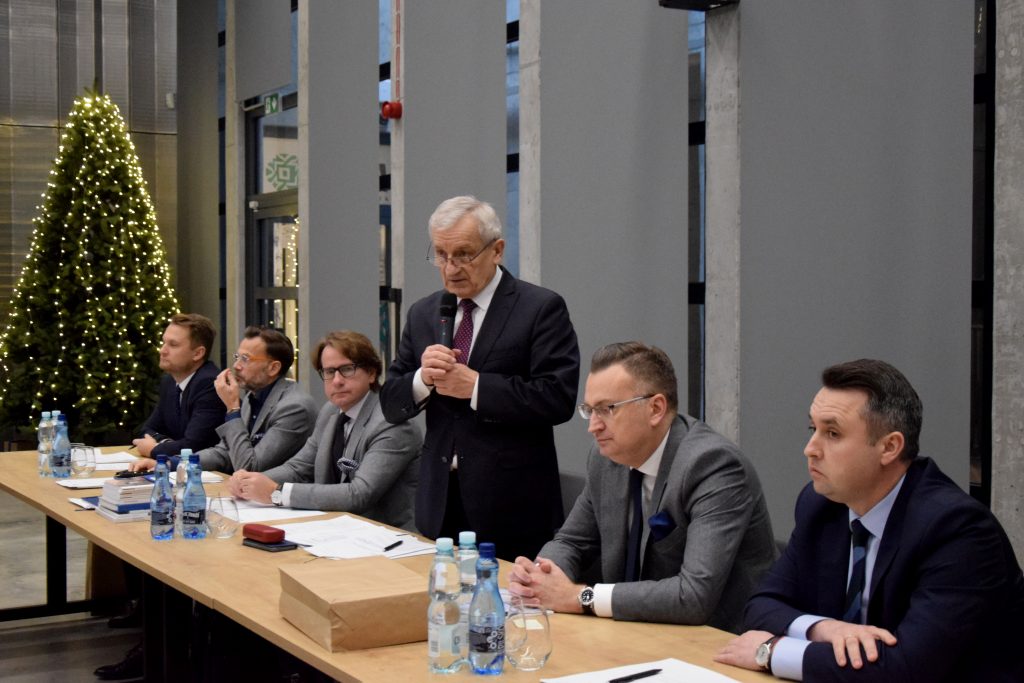 Mężczyzna w garniturze i krawacie przemawia przy mikrofonie, pięciu innych mężczyzn w garniturach siedzi przy stole. Na stole stoją butelki wody, szklanki i dokumenty, w tle choinka ozdobiona lampkami.