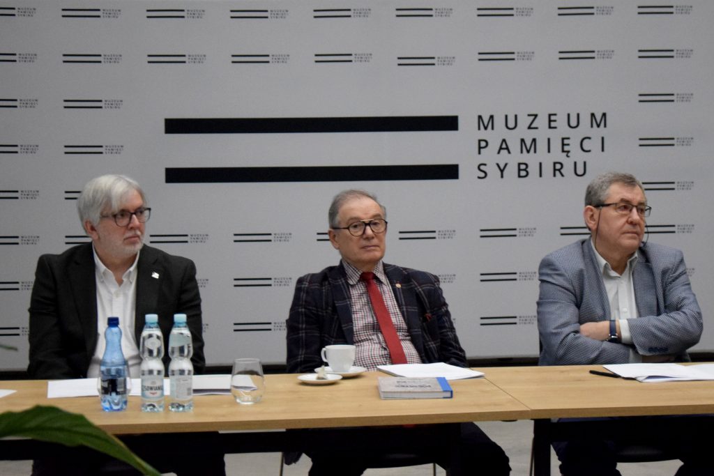 Trzech mężczyzn w garniturach siedzi przy stole. Na stole znajdują się butelki wody, notatki i książka. W tle logo Muzeum Pamięci Sybiru na białej ściance.
