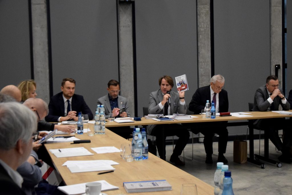 Kilkanaście osób w garniturach siedzi przy stołach, na których znajdują się butelki wody, notatki i dokumenty. Prof. Wojciech Śleszyński, dyrektor Muzeum Pamięci Sybiru, trzyma książkę i przemawia przez mikrofon.