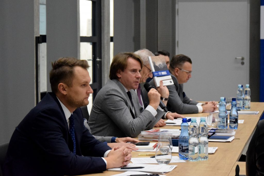 Kilka osób w garniturach siedzi przy stole, na którym znajdują się butelki wody, notatki i dokumenty. Prof. Wojciech Śleszyński, dyrektor Muzeum Pamięci Sybiru, trzyma książkę i przemawia przez mikrofon.