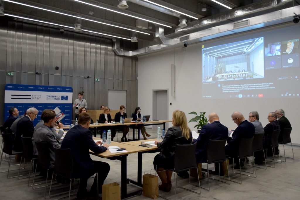 Kilkanaście osób w garniturach siedzi przy stołach, na których znajdują się butelki wody i notatki. Na ekranie wyświetlane są osoby, biorące udział w spotkaniu online. W tle logo Muzeum Pamięci Sybiru.