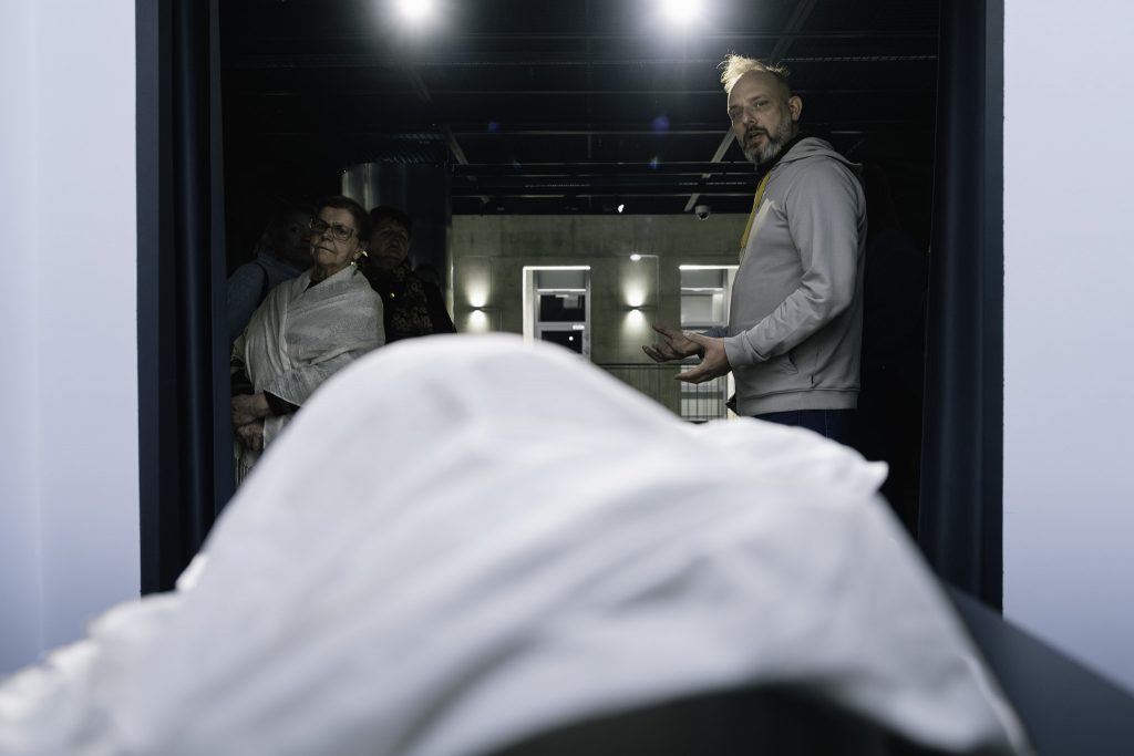 Na pierwszym planie znajduje się duży, biały przedmiot, który jest jedną z prac wystawy czasowej "Bezkres Sybiru". Po lewej stronie widać kilka osób, które stoją i przyglądają się. Obok nich stoi kurator wystawy.