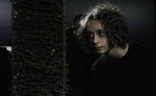 Młody chłopak z długimi, kręconymi włosami patrzy w bok z zaciekawieniem. Na pierwszym planie widoczna jest jedna z prac wystawy czasowej "Bezkres Sybiru".