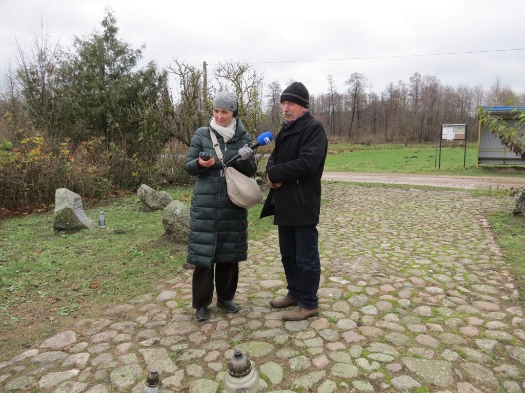 Dziennikarka trzyma mikrofon skierowany w stronę rozmówcy, w drugiej ręce trzyma dyktafon, na uszach ma słuchawki. Rozmówcą jest starszy mężczyzna.