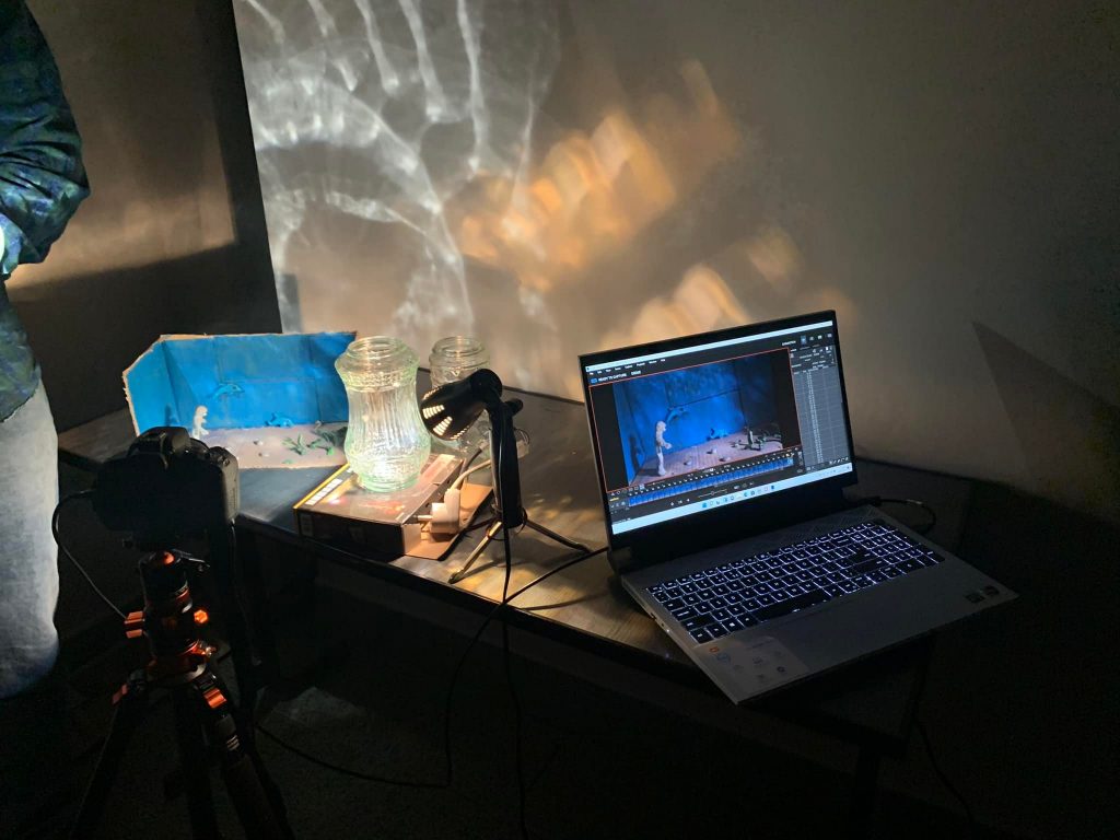 Na stole znajduje się laptop, na którym wyświetlany jest program do edycji wideo lub animacji, pokazujący scenę z postacią na niebieskim tle. Obok laptopa jest kamera na statywie skierowana na dwa szklane pojemniki, które są oświetlone od tyłu, tworząc efekt projekcji na ścianie. Na stole znajduje się również niebieska scenka z figurkami, prawdopodobnie używana do tworzenia animacji poklatkowej.