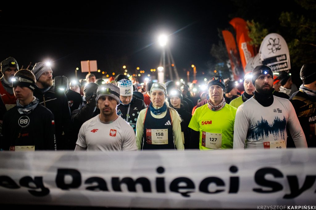 Grupa osób ubrana na sportowo, z czołówkami czeka na start.