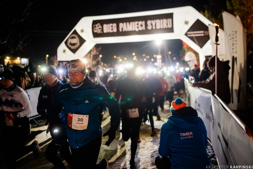 Grupa biegaczy uczestniczących w nocnym biegu po lesie. Biegacze są ubrani w odblaskowe stroje i mają czołówki. Każdy z nich ma numer startowy przypięty do ubrania. W tle napis Bieg Pamięci Sybiru.