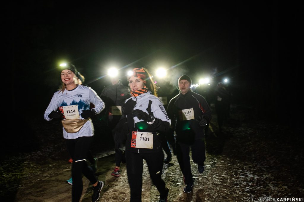 Grupa biegaczy uczestniczących w nocnym biegu po lesie. Biegacze są ubrani w odblaskowe stroje i mają czołówki. Każdy z nich ma numer startowy przypięty do ubrania.