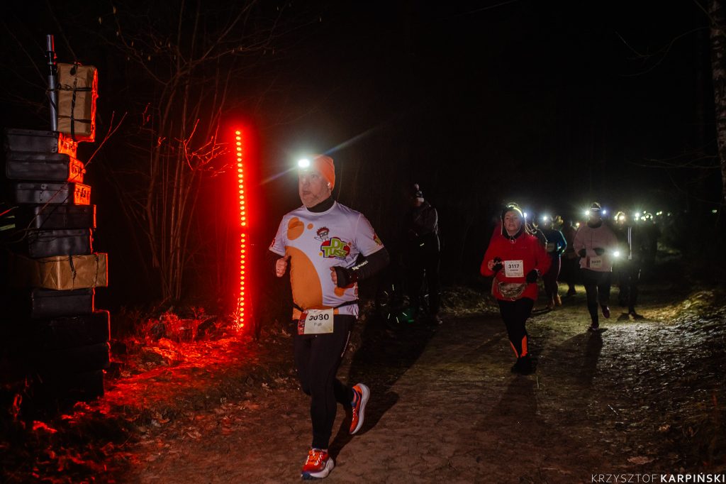 Grupa biegaczy uczestniczących w nocnym biegu po lesie. Oświetlone na czerwono walizki podróżne stanowią instalację artystyczną. Biegacze mają czołówki. W tle widać więcej uczestników.