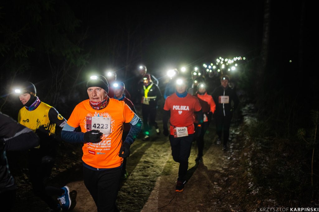 Duża grupa biegaczy uczestniczących w nocnym biegu po lesie. Biegacze są ubrani w odblaskowe stroje i mają czołówki. Każdy z nich ma numer startowy przypięty do ubrania.