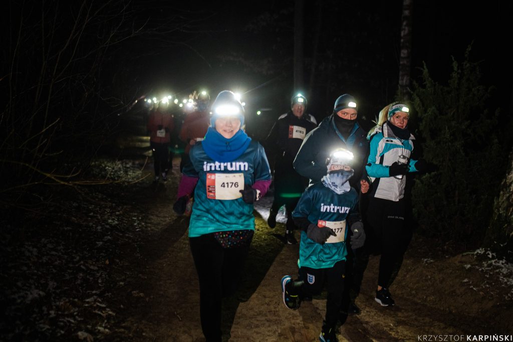 Grupa biegaczy uczestniczących w nocnym biegu po lesie. Biegacze są ubrani w odblaskowe stroje i mają czołówki. Każdy z nich ma numer startowy przypięty do ubrania.